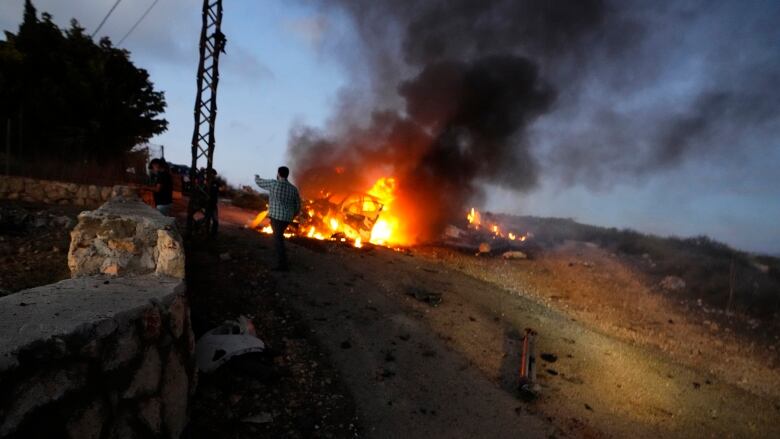 A man stands in front of a burning car.