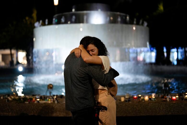 Two people embrace at night in front of a fountain.