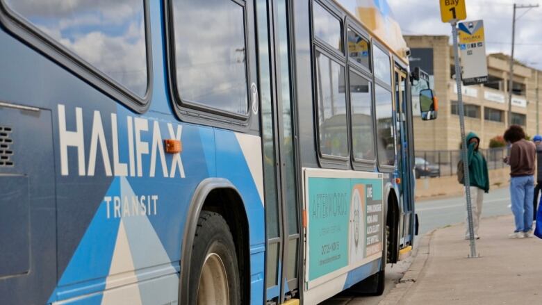 The side of a bus is shown with people waiting on the platform in the background