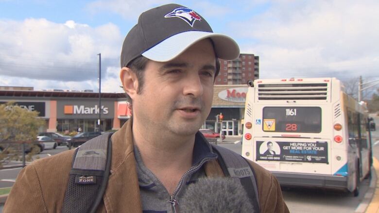 A man in a tan jacket and wearing a baseball cap speaks to the camera.