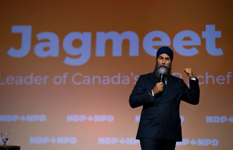 A bearded man, wearing a turban and a dark suit, gestures as he speaks into a microphone.