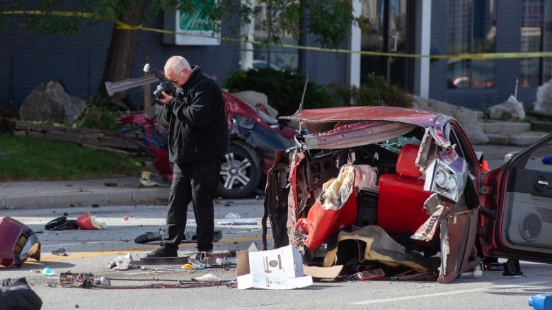 A man takes photos of a red vehicle that is crashed and in two pieces on the street. Detritus is all around him, including broken glass and unidentifiable contents from the vehicle