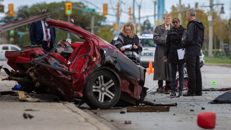 Investigators look at half of a red vehicle that has been destroyed in a crash and is angling upward onto the sidewalk