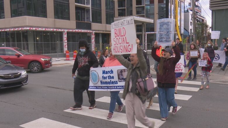 A small group of people carry signs calling for more housing while walking on a crosswalk.