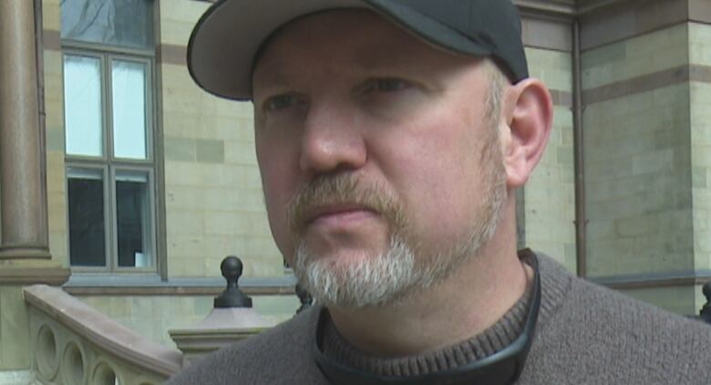 A man in a grey woolen sweater speaks to the camera.