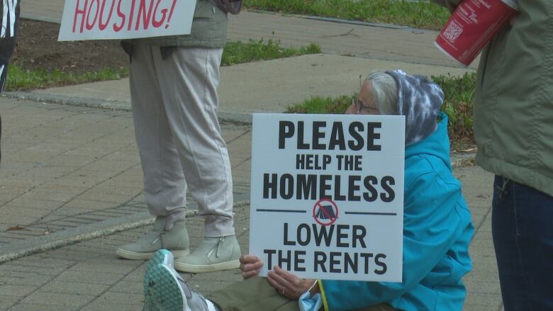 An elderly woman sits on the ground holding up a sign that says 