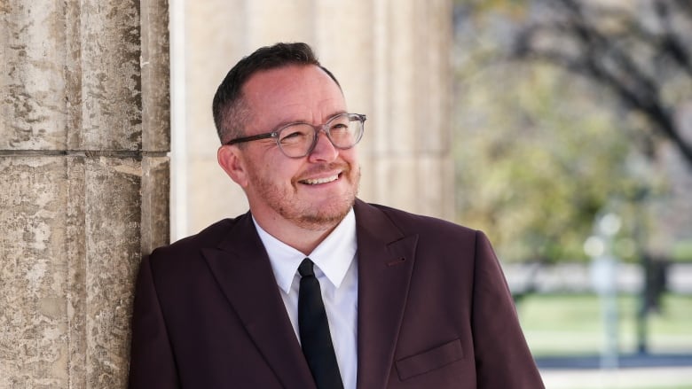 A person wearing a suit and tie smiles as he leans on a column attached to a building.