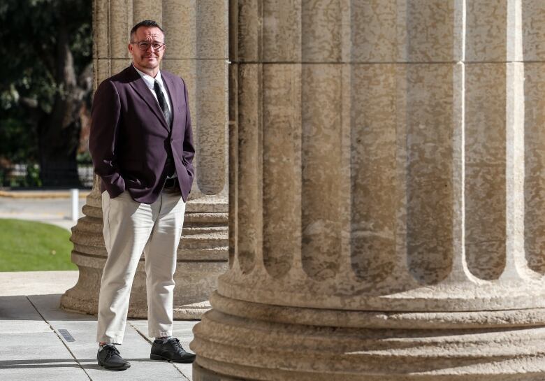 A person wearing a suit and tie stands next to a column attached to a building.