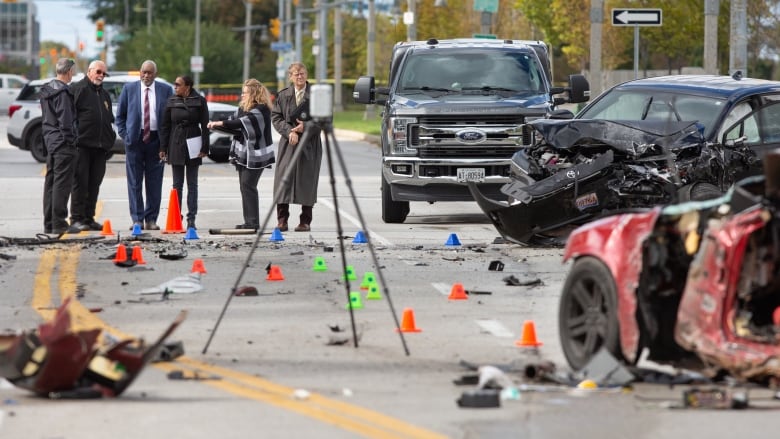 A group of investigators stand on the street in the middle of three vehicles, two of which are destroyed. There are remnants of glass and rubber around them