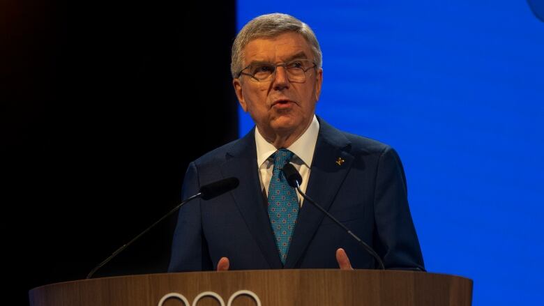 A man in glasses and a suit speaks at a podium that is adorned with the five Olympic rings.