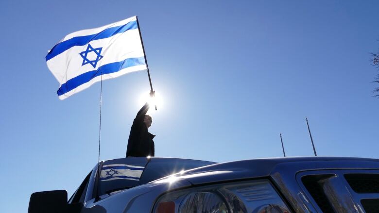 A person waves an Israeli flag from a vehicle, with the sun shining behind it.