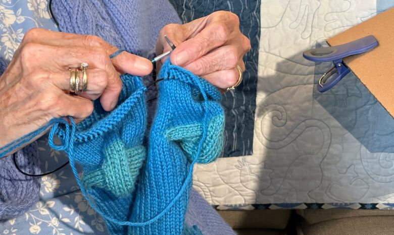 A pair of blue socks are shown close up with a woman's hands working the knitting needles to make them.