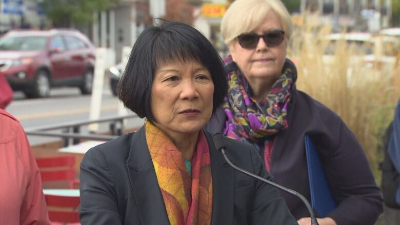 A middle-aged woman stands in front of a podium and microphone on a city sidewalk. Patio chairs are visible in the background. It's a grey, fall day.