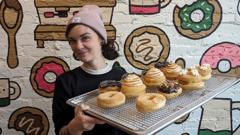 Woman wearing pink hat holds a tray of decorated donuts in front of a wall with cartoon-drawn donuts.