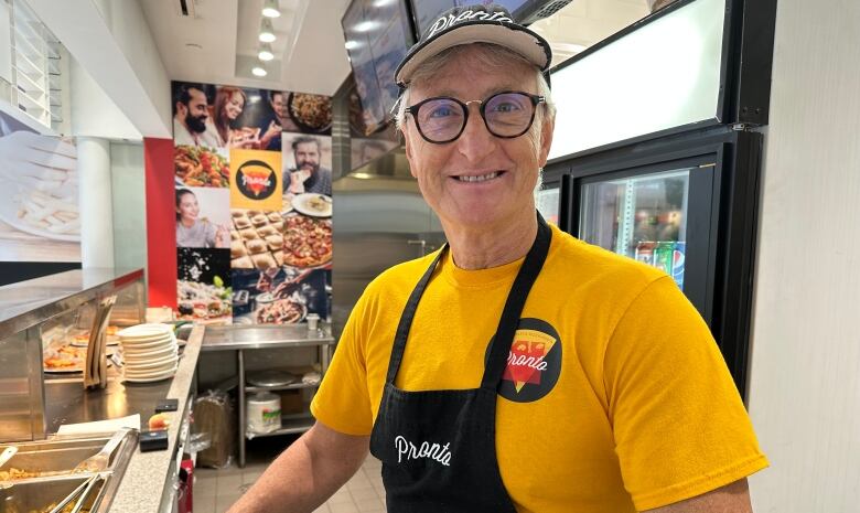 A man in a yellow shirt and apron smiles behind the counter of a pizza and pasta restaurant.