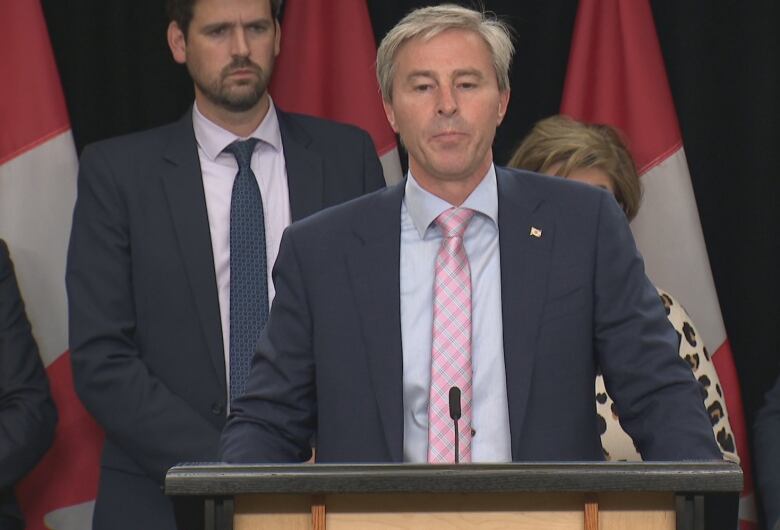 A man in suit and tie stands in front of Canadian flags.