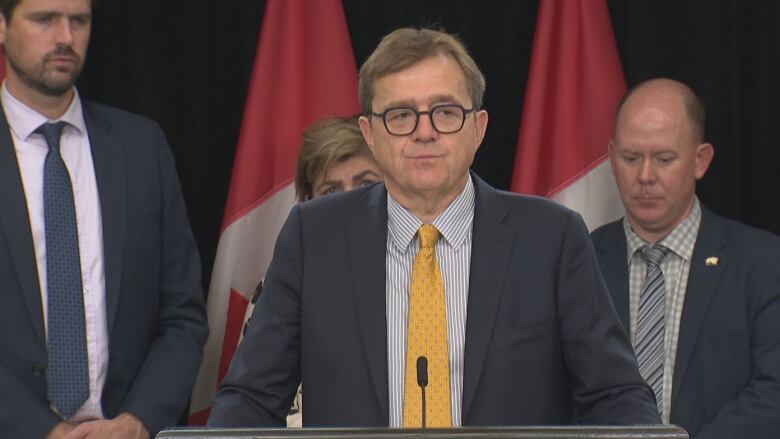 A man in a suit and tie and glasses stands in front of Canadian flags.