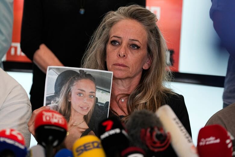 A woman who looks distressed holds up a picture of another, younger woman while seated in front of microphones.