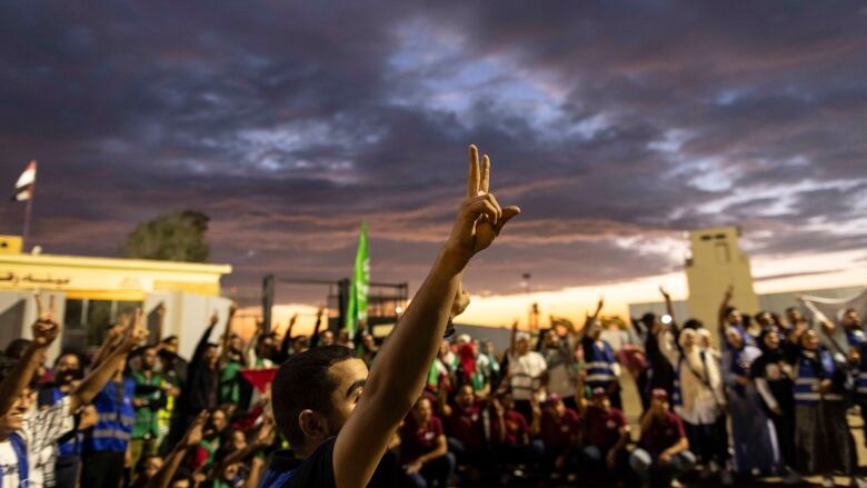 Early morning, a man flashes a peace sign with one hand surrounded by other people; he's part of volunteers and NGO staff on the Egypt side of the border crossing with Rafah, Gaza.