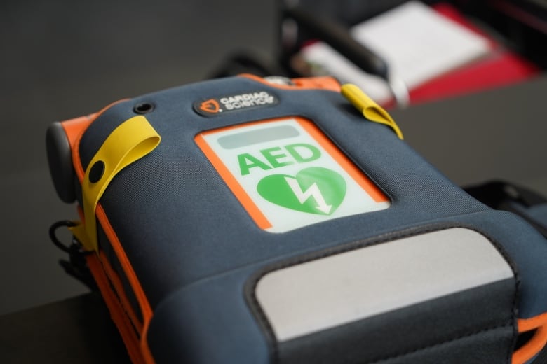 A grey and blue device is secured in an orange holder with yellow straps. Green letters on a grey background spell out AED above a green heart. In the heart is a white lightning bolt that ends in an arrow point.