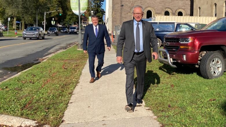 Two men in suits walk down a sidewalk. Traffic is visible on the road beside them.