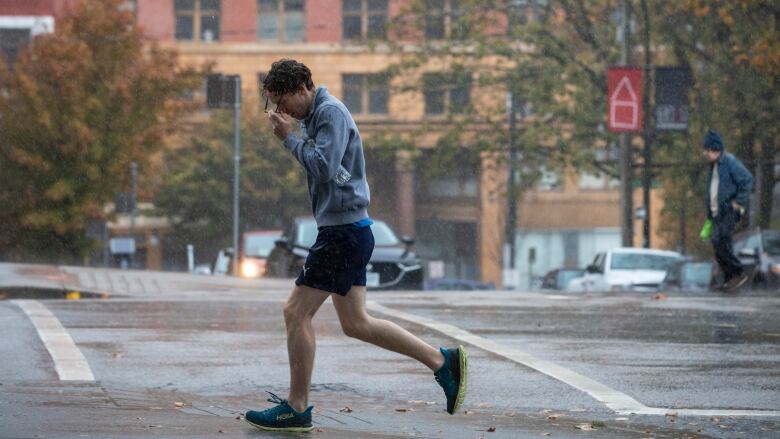 A wet-looking person in running gear holds his glasses in the rain as he steps onto the sidewalk from an intersection in Vancouver. 