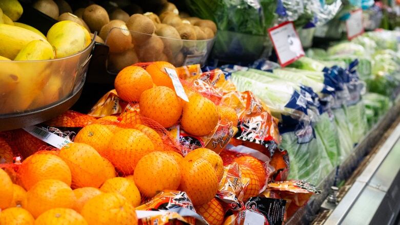 A view of produce on grocery store shelves, with bags of oranges in the foreground, and bags of lettuce further along the aisle.