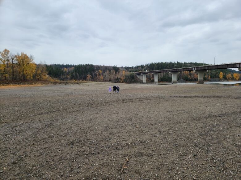 A family of three walks on a gravel river bottom amidst low water levels, with a railway bridge in the background.