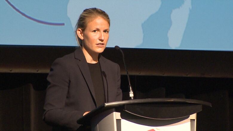 A portrait of a woman wearing a black blazer and black top, with her hair pulled back, standing at a podium, speaking into a microphone.