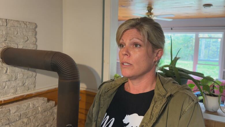 A woman with blonde hair wearing a green jacket stands next to the pipe from a wood stove. 