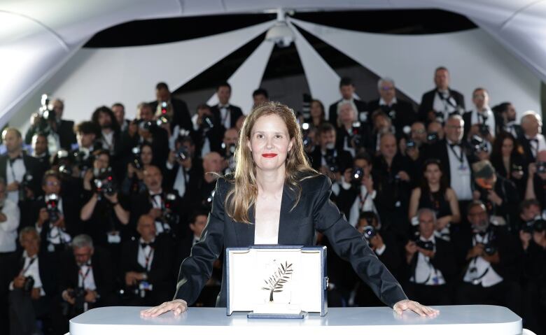 A woman stands in front of a desk, on which a plaque sits. Behind her are is a crowd of photographers taking her photo. 