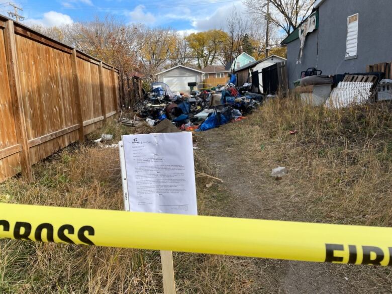 A white City of Regina notice is posted behind a yellow fire scene tape. Further back in the empty lot are the remains of a homeless encampment after a fire. 