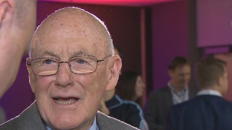 A portrait of an older man wearing a suit and tie, speaking to a younger man in a suit, whose back is to the camera.