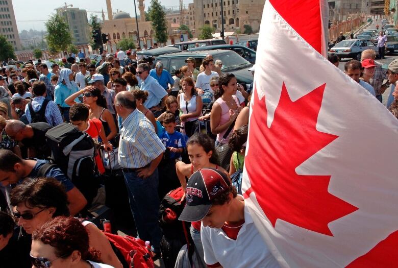 A Canadian flag is held up, on the right side of the image, above a large crowd of people stand outside.