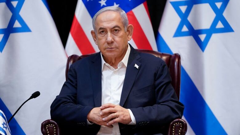 Israeli Prime Minister Benjamin Netanyahu sits in a  chair in front of several Israel flags, answering questions.