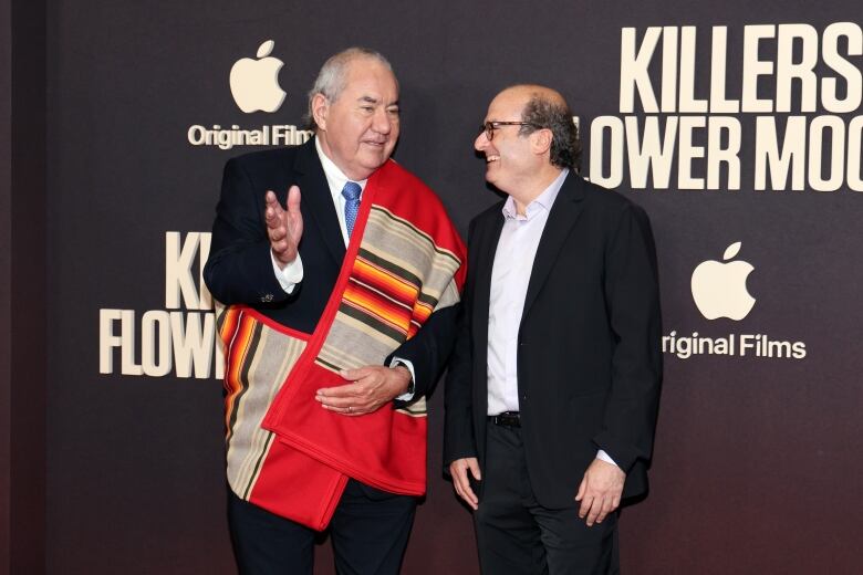 Two men, one wearing a traditional Osage blanket, smile and talk in front of a backdrop.