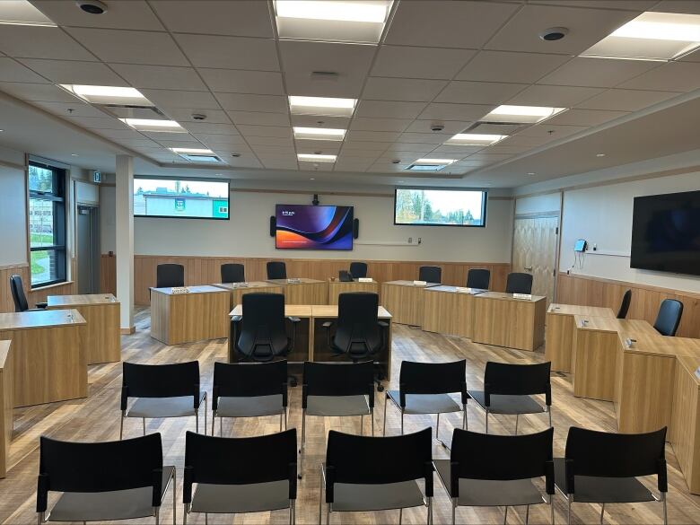 Three television monitors sit on the back wall of a city hall council chambers with chairs and desks arranged in a semi circle.