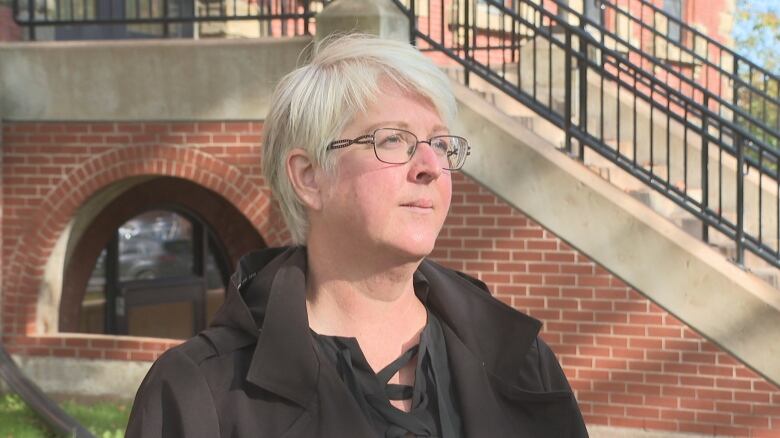 Woman in glasses with short white hair stands outdoors in front of a brick staircase.