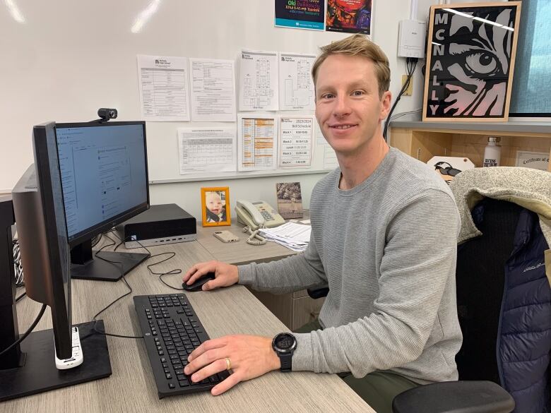 Man in grey sweater sitting at an office desk in front of a a computer.