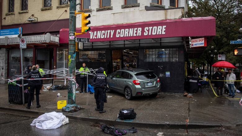 Police officers set up a shelter in front of the Vancity Corner Store, where a silver car is crashed into the entrance.