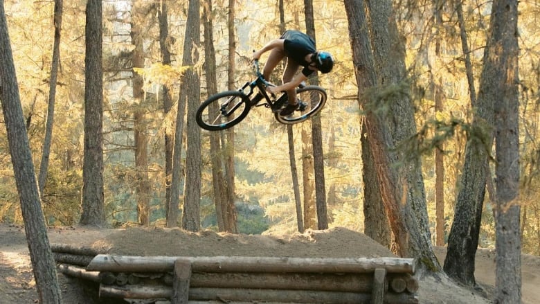 A man on a bike spins in the air while launching off a dirt jump on a track in a forested area with dappled sunshine.