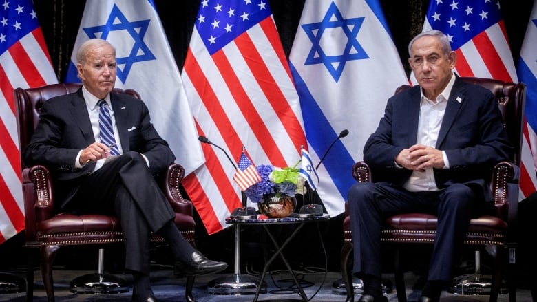 Two grey haired men in suits sit in front of the flags of Israel and the U.S.