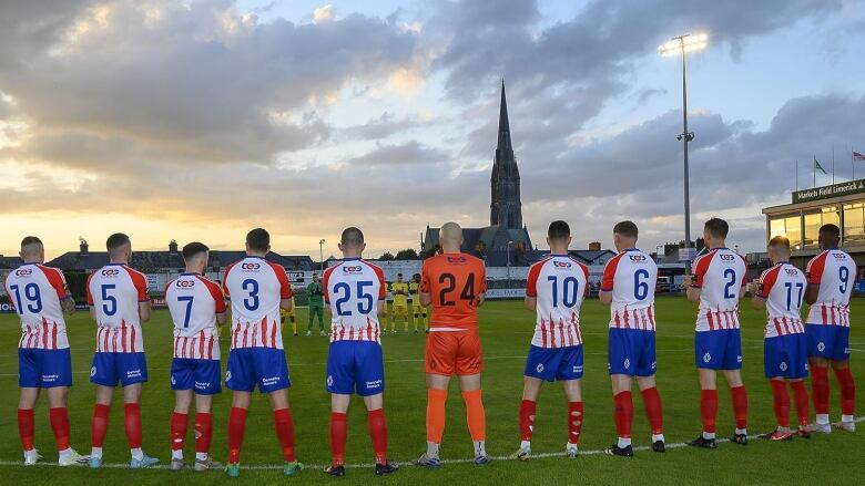 Male soccer players stand with their backs to the camera at Markets Field Stadium in Limerick, Ireland.