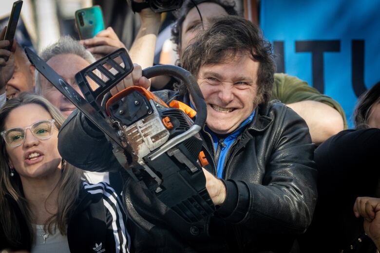 A man in a leather jacket stands in a crowd, holding a chainsaw.