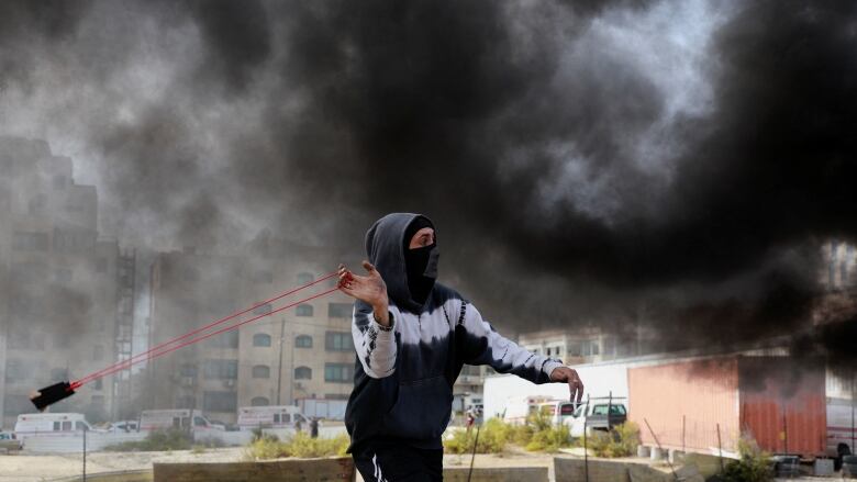 A person with a head covering and a mask uses a slingshot-type device while a massive cloud of black smoke is seen behind them.