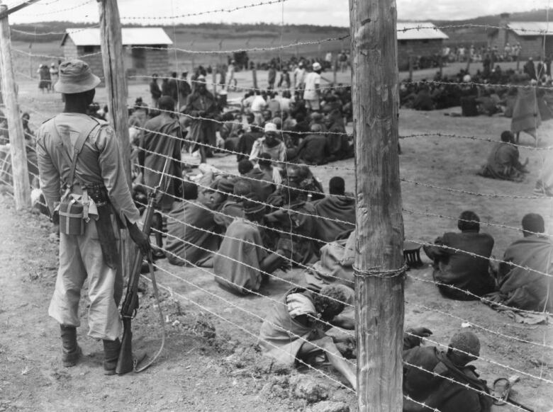 People sit on the ground inside an enclosure surrounded by barbed wire as a soldier looks at them.