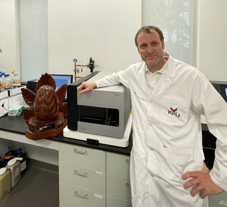 A man in a white lab coat stands in a laboratory with his arm resting on a small cube-shaped machine.