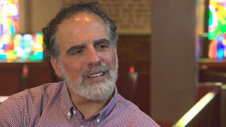 A man with dark grey hair and a light grey beard is pictured speaking.