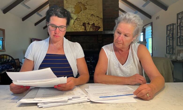 Two white woman sit at a marble table inside a home and look through documents on the table. 