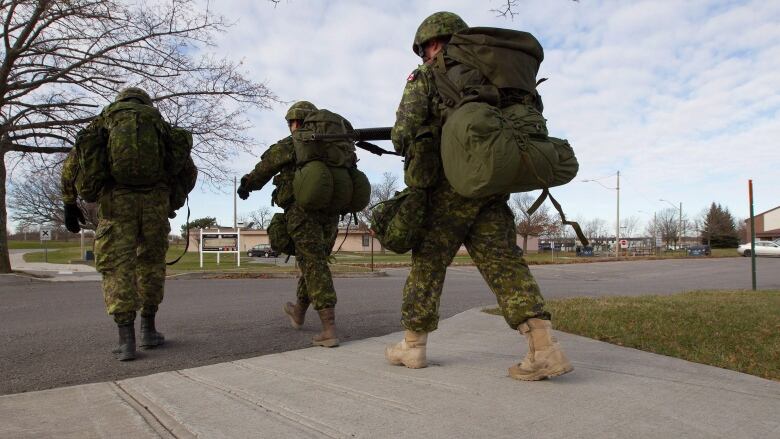 Three soldiers, laden with gear, march across the grounds of a military base. 